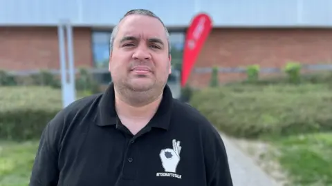 Michael Chapplow, standing in front of the Beacon of Light in Sunderland. He is wearing a black polo shirt with the logo for Andy's Man Club. The logo for the club is a hand making the "ok" sign with writing which reads # It's Okay To Talk. He has stubble and very short dark hair flecked with grey.