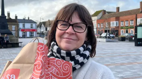 Tom Jackson/BBC Nikki Derrick is standing in the middle of a market square. She has short brown hair and is wearing black framed glasses, and black and white striped scarf and white jumper.