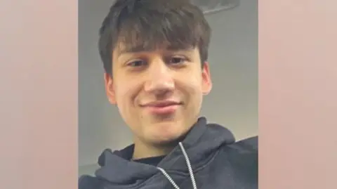 A portrait selfie of a young man wearing a black hoodie. He has brown hair with a fringe over his forehead and brown eyes. 