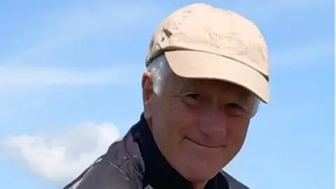 Family handout  Richard is wearing a cream hat.
He is smiling at the camera with blue clouds behind him and one fluffy white cloud. 
He is wearing a black raincoat and the photo is cropped from the shoulders up.