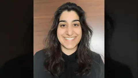 Tasmiyah Manan A woman with long dark hair is shown in a portrait against a smooth wooden background. She’s wearing a black collared shirt and looking directly at the camera.