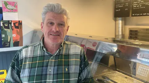 A man with grey hair, wearing a green checked shirt, smiles at the camera as he leans against a chip shop counter. 