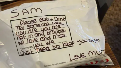 A parcel. On the front is written: "Sam, please eat and drink. Tell someone where you are. You are not in any trouble. We love and miss you. We just need to keep you safe. Love mum." 