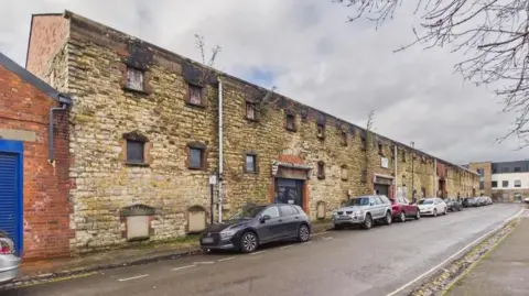 The outside of an old music venue. It is a stone brick, warehouse-style building. It looks quite run down, with boarded up windows. There are cars parked alongside it.