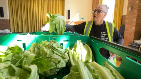 Gail Seamark is picking up a cabbage from a green box full of the vegetables and putting it into a white plastic bag. Gail has her brown hair, which is tied up, and is wearing black-rimmed glasses. She is wearing a black shirt and a yellow high-vis jacket.