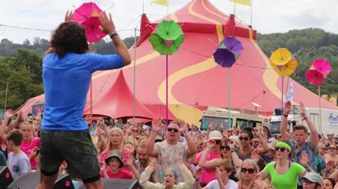 Joe Wicks wearing a blue t-shirt exercises whilst looking at the crowd who are facing him in front of a yellow and red festival tent