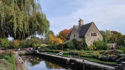 Dawnie61 A house next to a canal under a mostly cloudy sky. Trees can be seen in the background with orange and yellow autumnal colours