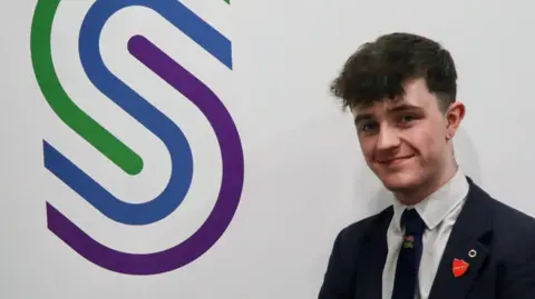 Shimna Integrated College A boy with dark hair in a navy blue school uniform. He is standing in front of a white wall which has the school logo to his left.