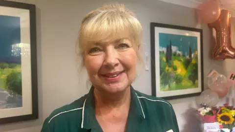 Jane Rogers wearing a green nurses uniform and name tag. She has blonde hair which is tied up, with a thick fringe. She is smiling at the camera, and behind her you can see artwork on the wall and a table full of flowers and cards. There are also rose gold mylar balloons which say '100'.