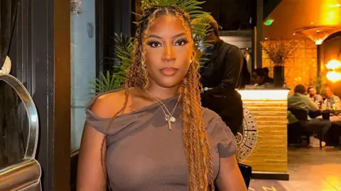 Khiefa Gabbidon A woman with long strands of plaited golden hair, wearing a necklace and a brown top, sits at a table in a restaurant.