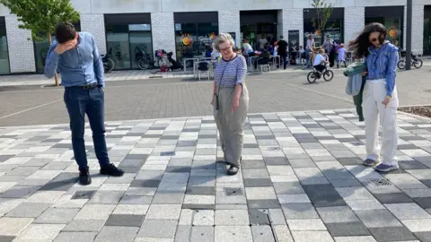 Emma Howgego/BBC A man and two women point to small squares in the pavement which look like drain covers. They are standing in front of a cafe. 