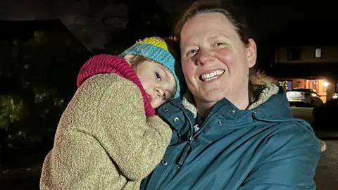 A woman wearing a blue coat stands on a dark street smiling at the camera. She is holding a small child wearing a brown coat and a woolly hat who is leaning on her shoulder.