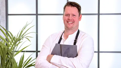 Thom Bateman, a man wearing a white shirt and a grey apron, is standing with his arms folded. He is smiling while posing for the photograph. He is standing against a set of illuminated white panels. There is a plant next to him.