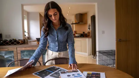 Sasha-Jay looking at the multiple A4 print outs of the fake accounts which are layed on her kitchen table. Her right hand is holding onto a pile of the images, while her left hand is touching one of the images on the table. 