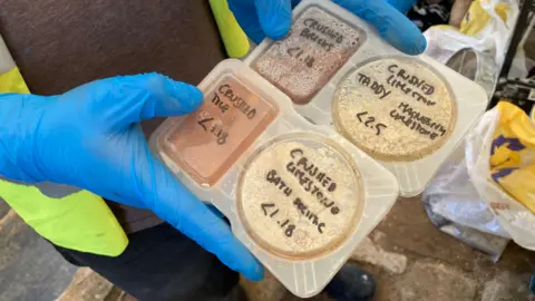 BBC / Jack Hadaway-Weller A close-up shows gloved hands holding a plastic tray filled with labelled material samples. Each compartment contains crushed or powdered substances with handwritten notes such as “crushed tile,” “crushed limestone masonry,” and similar labels. The scene appears to be part of a restoration or construction project, focusing on analysing building materials.