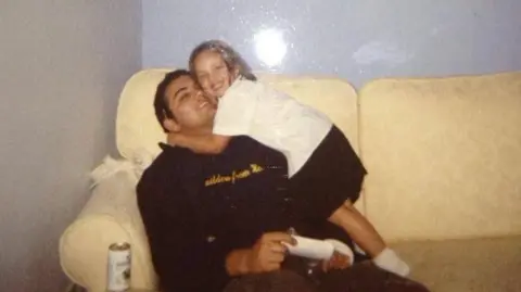 Chloe Cooper An old image of Chloe hugging her dad who is sat on a yellow sofa. He wears a black hoodie, while Chloe is in school uniform. Both are smiling looking at the camera.
