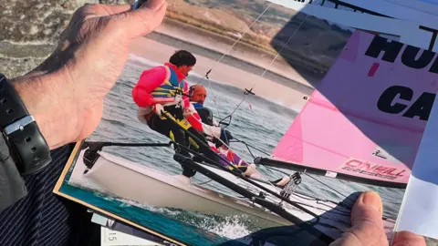 Close-up of hands holding a photograph of two people sailing a small catamaran with a bright pink sail. The photo is partially inside a folder, and the background shows a rocky surface.