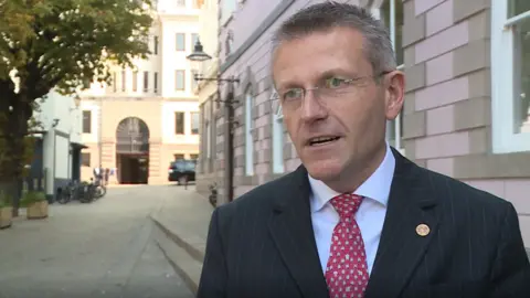 Philip Ozouf in a black suit, white shirt and red tie, standing outside the States building in St.Helier, Jersey