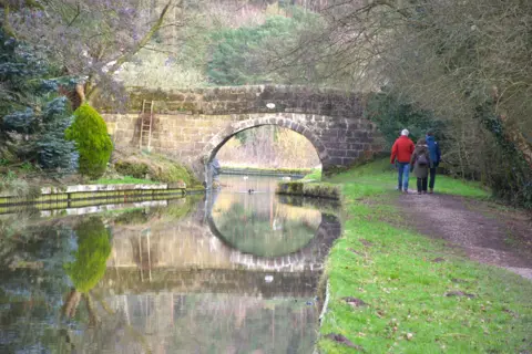 BBC Weather Watchers/RKP A brick canal bridge is reflected in its water as three people, two adults and a child, walk away from us in the distance along a canal towpath.