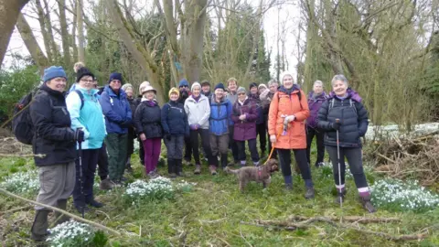 Sid Mill This picture shows a group of approximately 19 people with their warm hats and coats on. Two either side of the photo have walking poles. One person has a small brown dog on an orange lead that matches her orange coat. They are standing in front of woodland on grass with large clumps of snowdrop flowers dotted around them.