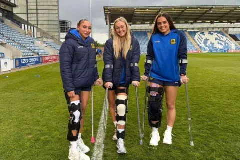 Hashtag United Football Club Three women in blue football training kit, on crutches and leg braces, while standing on a football pitch. The blue seating of Colchester United's stadium stands are behind them.