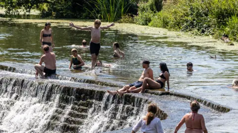 A group of wild swimmers in the River Avon at Warleigh Weir. Some people are swimming while others are standing in the water or sitting by the edge of the weir. One man has his arms outstretched as he stands in the water.