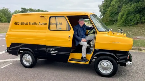 BBC BBC business correspondent Theo Leggett sits with one hand on the wheel of a stationary yellow Ford Transit - the oldest one still in existence, which was built in 1965. On its side are the words GEC-Elliott Traffic Automation Ltd. He is smiling wearing a cap and a blue shirt and jacket and light grey trousers.