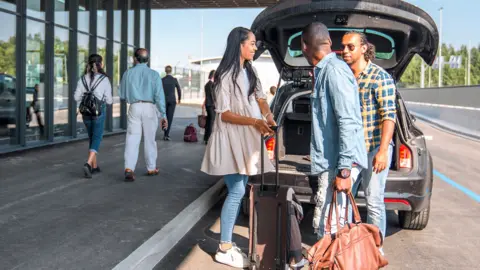 Stock photo shows a group of people removing luggage from the boot of a car as it pulls up to an airport with other passengers walking past.