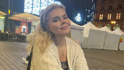 Anya Roberts Anya Roberts smiles at the camera, in a market square. She has blonde wavy hair and a white headband, and wears a white cardigan and a patterned top.