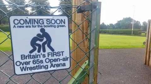 Carp A joke sign reads: Coming soon. A new era for the bowling green. Britain's first over 65s open-air Wrestling Arena