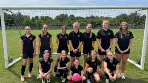 A photo of 12 members of a female football team, the front row sitting and the back row standing, in front of a goal. They are wearing black and yellow football kids. The girl in the middle of the front row is holding a pink football. They are all looking at the camera and smiling.