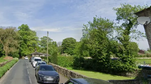 Google A queue of parked cars along a narrow, rural road. A pavement and dry stone wall are to the side of the cars. Some trees and shrubs are in the background.