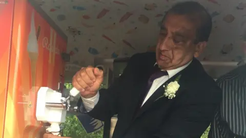 Supplied Dipak Davda wearing a black suit and tie serving ice-cream into a cup