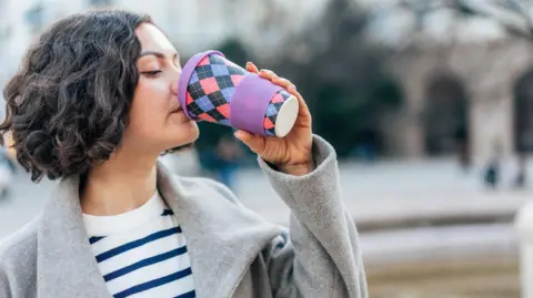 Young woman with dark hair in a short bob wearing a grey wool coat over a blue and white stripped top drinks from a reusable cup