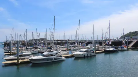 The marina is full of small yachts and there are three speedboats moored up in the foreground. It is a bright and sunny day.