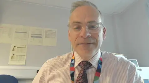 A man wearing a white and light pink striped top and black tie with pink flowers. He is wearing glasses. He is wearing a colourful lanyard. In the background is a white wall with pieces of paper stuck to it. 