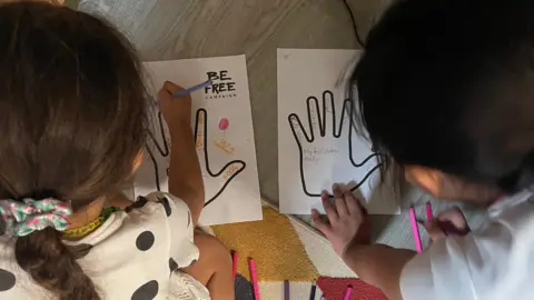 Be Free Campaign A picture of two young children completing a "memory hand" exercise. The picture is taken over their shoulder and shows them writing positive memories inside the outline of a hand.