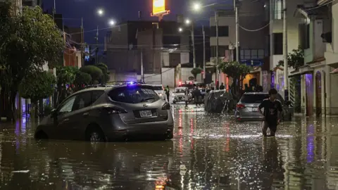 Getty Images A man walks next to a car in the middle of a flood caused by heavy rains in the Yanahuara district of Arequipa