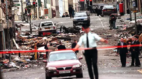 Red tape is taped across a street in the aftermath of a bomb. A police officer is standing in the foreground with his arms up. Behind him, rubble and debris cover the street and road. Cars are damaged with glass blown out.