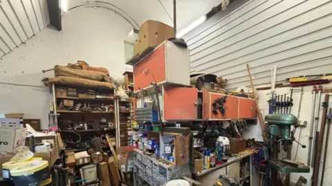 Brian Farmer/BBC A room with a high white ceiling and white panelled walls, lined with shelves and cupboards full of tools and boxes.