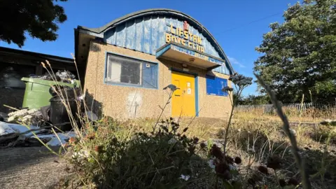 The Lifstan Boys Club building, surrounded by overgrown grass and weeds, with an overflowing green bin to the side. The building has a pebble-dashed front with two blue-framed windows and a yellow front door. The upper half of the building is a domed curved roof with blue metal fronting and the words Lifstan Boys Club in yellow writing.