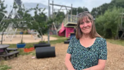 Alison Woods stands in the Meriden Adventure Playground with a huge climbing frame and climbing towers behind her. She has brown hair with glasses perched on her head and wears a floral dress.
