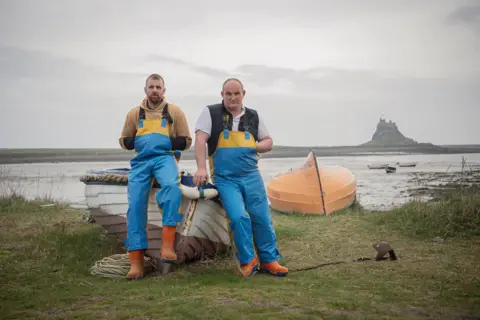 Jennifer Charlton Two fishermen in blue overalls lean either side of the bow of a small fishing boat, grounded on grass beside the sea, with a small castle across the water in the distance.