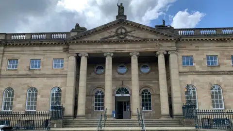 BBC/Oli Constable The front of a neoclassical building constructed from pale beige stone. The building is symmetrical, with a grand central entrance approached by a wide flight of stone steps. Six large columns support a projecting portico and triangular pediment above the entrance. Black wrought-iron railings line the front boundary and frame gateways on either side of the entrance.