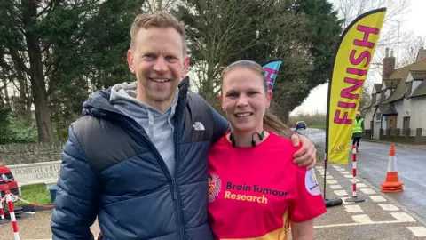 A brother and sister - aged around late 30s early 40s - smile as they put their arm around each other. They are stood at a closed off junction at the finish line of a road race in a Gloucestershire village on a winter's day. The man, who has short brown hair, is wearing a navy and black puffer jacket with a grey hoodie underneath and the woman is wearing a bright red Brain Tumour Research top. She has her brown hair slicked back into a ponytail.
