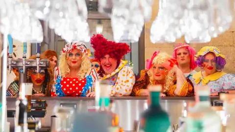 Hugh Hastings Six panto damges in bright costumes behind a bar with glasses above.