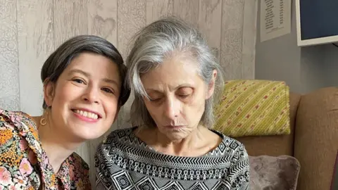 Sara Blagborough A woman with greying dark hair, wearing a flowery top, sits next to an elderly woman with grey hair, wearing a black and white patterned top.