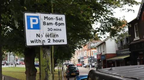 A parking restriction sign saying Mon-Sat 8am-6pm 30 mins next to a row of cars and shops in Harpenden