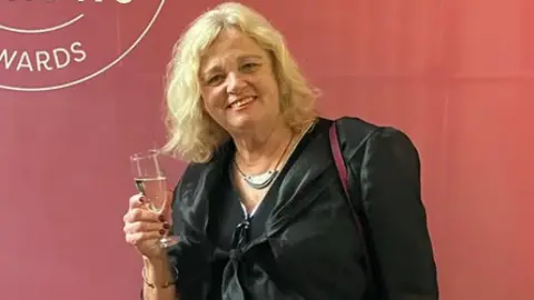 Helen Holland Helen holding a glass of proseccoin front of a red wall. She is dressed in a formal black dress and smiles at the camera. 