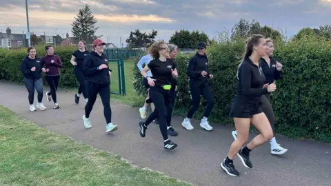 BBC / Kate Bradbrook Women runners mostly wearing black tops and leggings run along a path left to right, behind them is a park. The sky is cloudy approaching sunset.